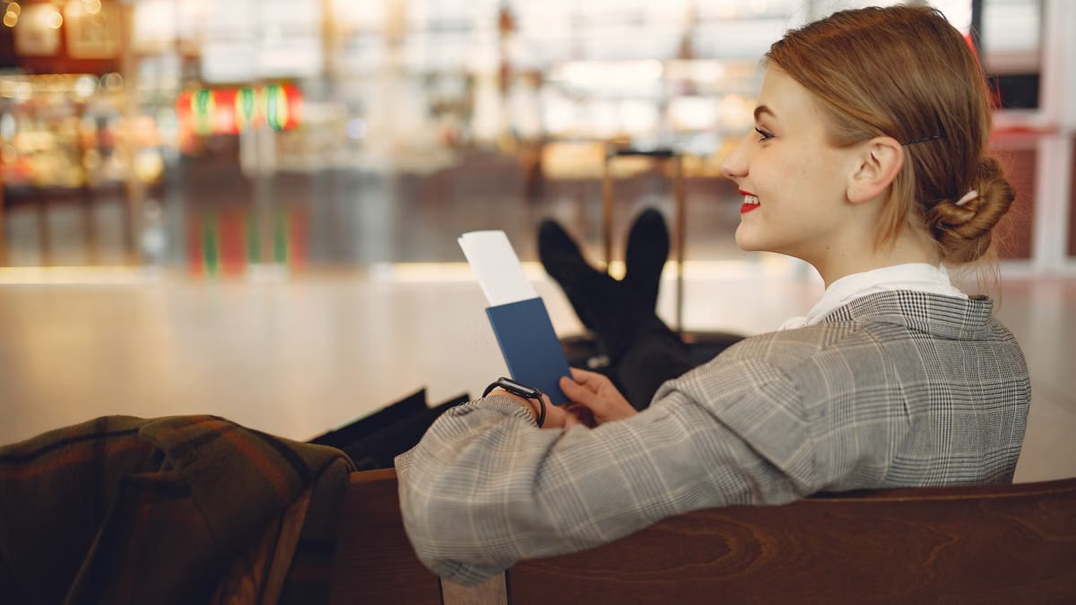 Side view of cheerful female student in checkered jacket smiling away while chilling in hall with outstretched legs and passport in hands